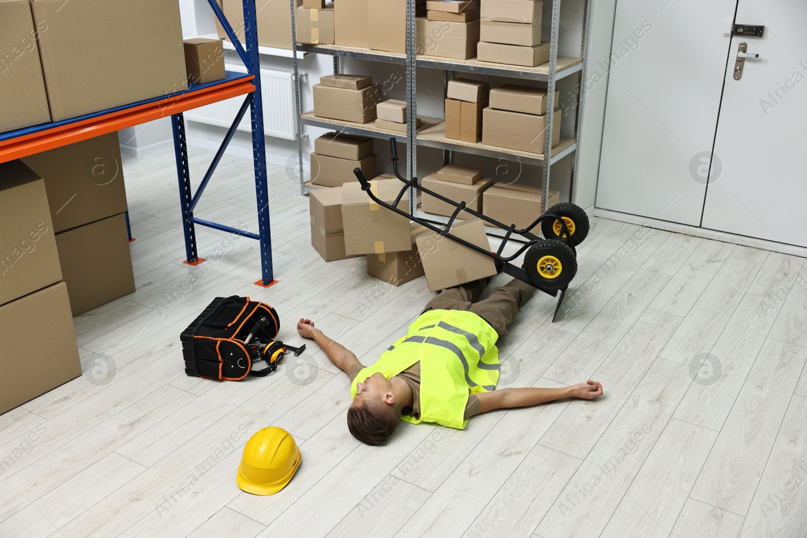 Photo of Accident at work. Unconscious man and overturned hand truck on floor in warehouse