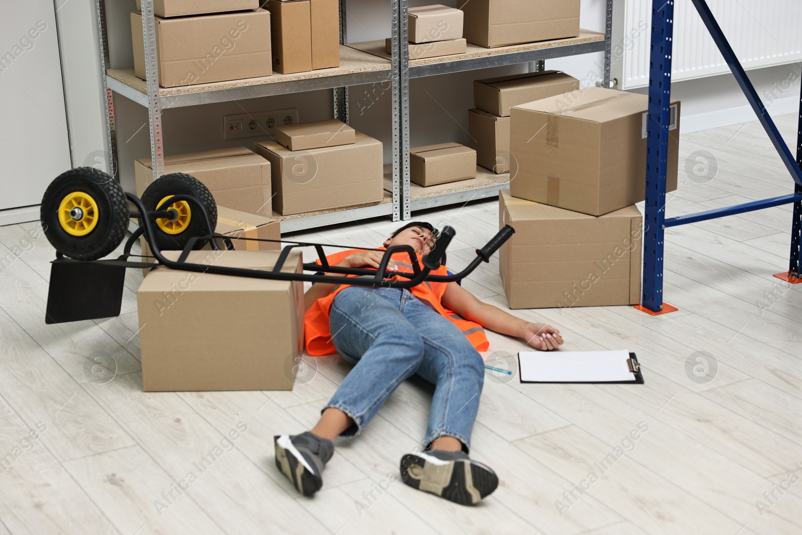 Photo of Accident at work. Unconscious woman and overturned hand truck on floor in warehouse