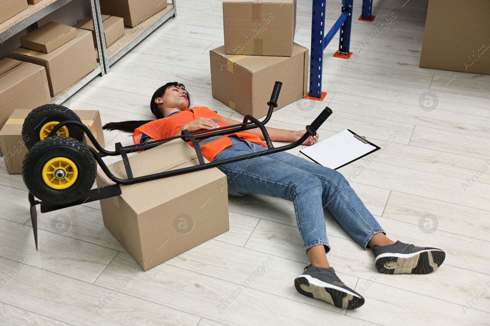 Photo of Accident at work. Unconscious woman and overturned hand truck on floor in warehouse