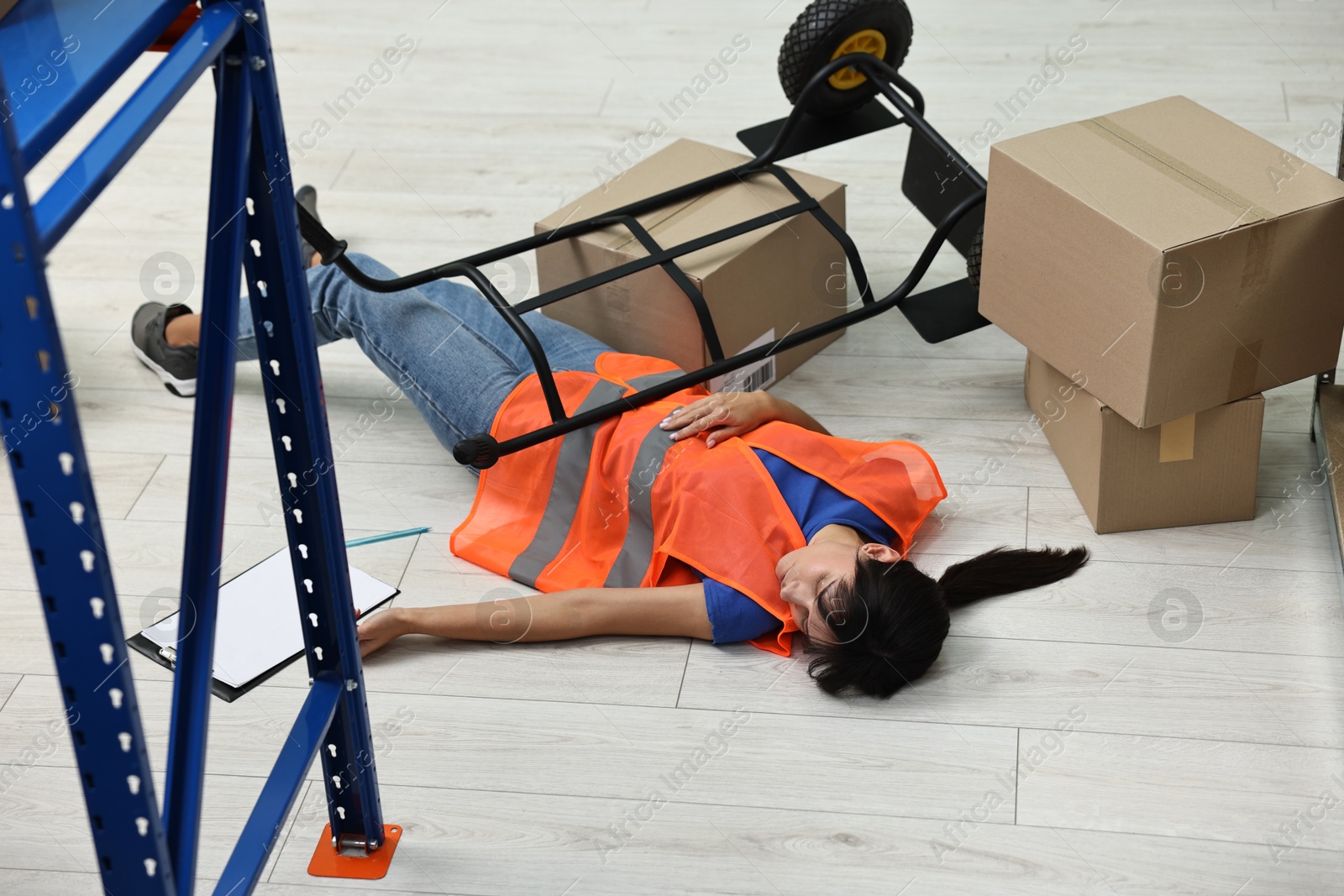 Photo of Accident at work. Unconscious woman and overturned hand truck on floor in warehouse