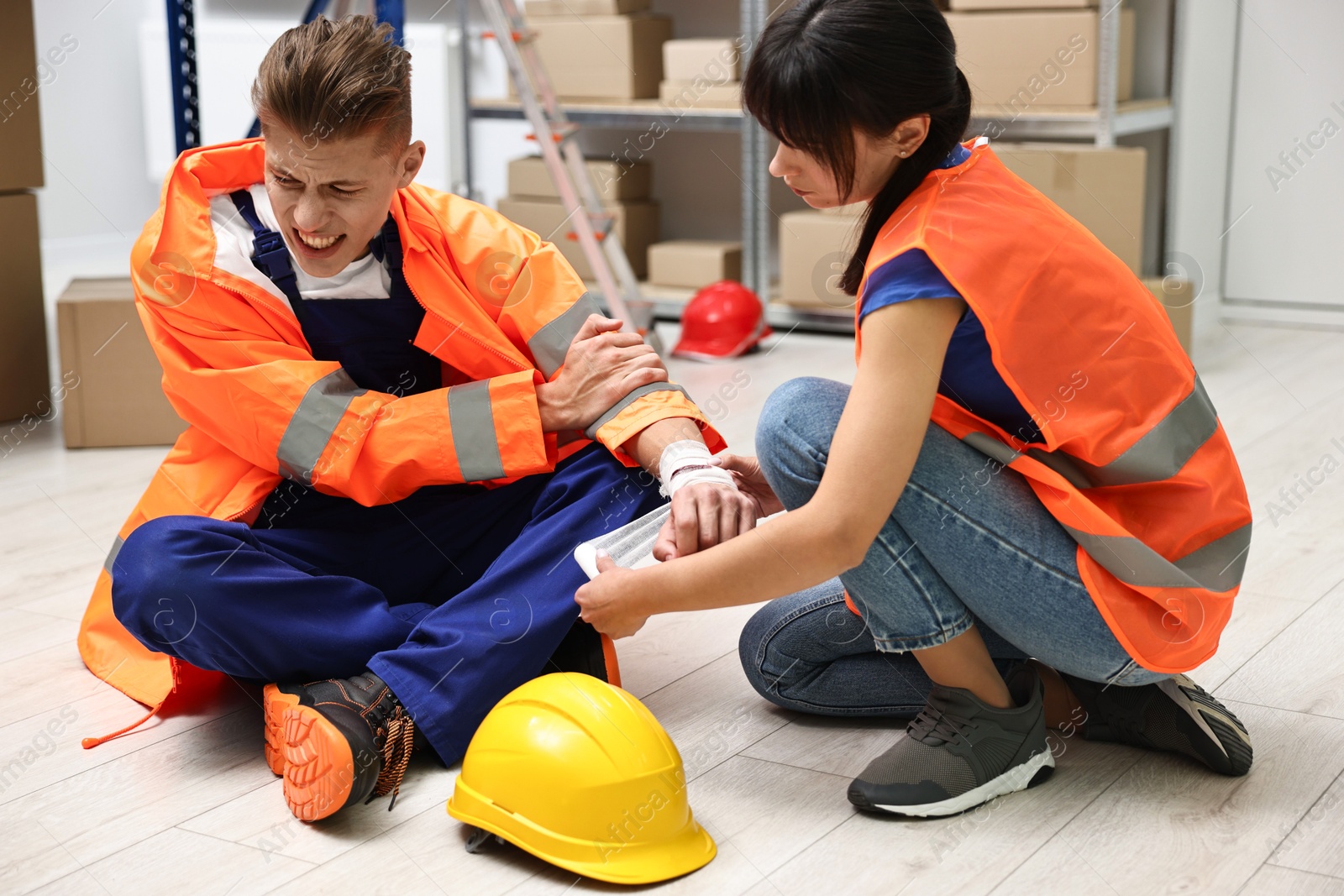 Photo of Accident at work. Woman putting bandage on her colleague's injured wrist in warehouse