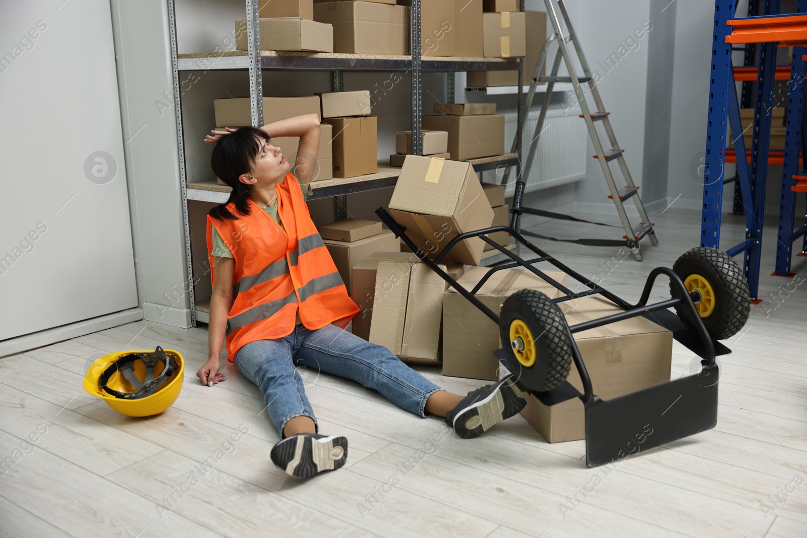 Photo of Accident at work. Injured woman and overturned hand truck on floor in warehouse