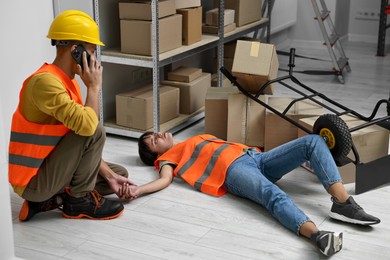 Photo of Accident at work. Man calling ambulance for his unconscious colleague in warehouse