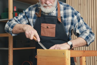 Photo of Relaxing hobby. Senior man repairing wooden stool with chisel in workshop, closeup