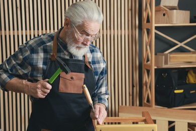 Relaxing hobby. Senior man repairing wooden stool with hammer and chisel in workshop