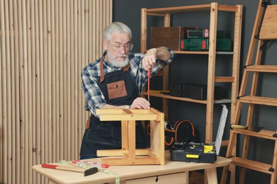 Photo of Relaxing hobby. Senior man repairing wooden stool with screwdriver in workshop