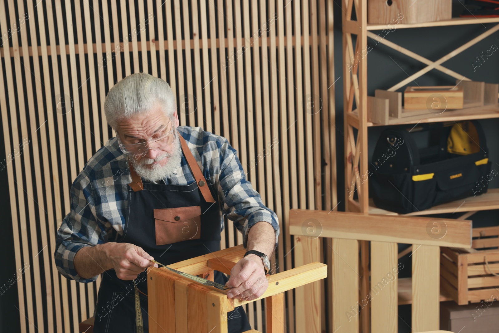 Photo of Relaxing hobby. Senior man measuring wooden stool with tape in workshop