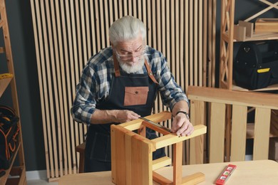 Photo of Relaxing hobby. Senior man measuring wooden stool with tape in workshop