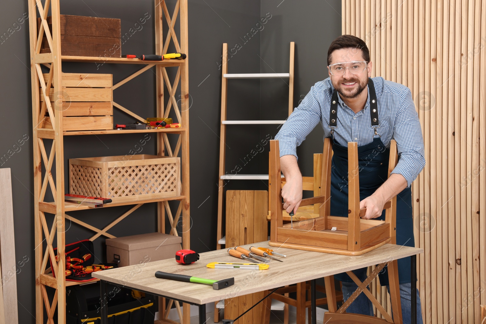 Photo of Relaxing hobby. Smiling man assembling wooden chair with screwdriver at table in workshop