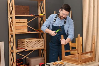 Photo of Relaxing hobby. Man repairing wooden chair with hammer at table in workshop