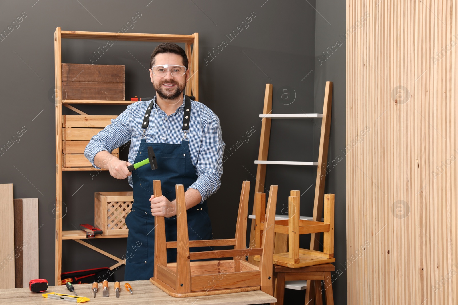 Photo of Relaxing hobby. Smiling man repairing wooden chair with hammer at table in workshop