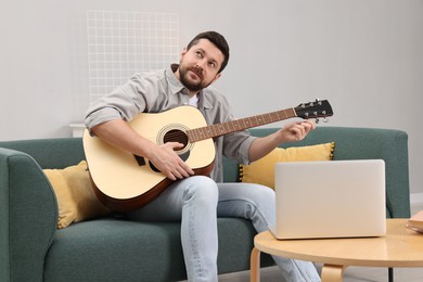 Photo of Relaxing hobby. Man playing guitar with online tutorial by laptop on sofa at home