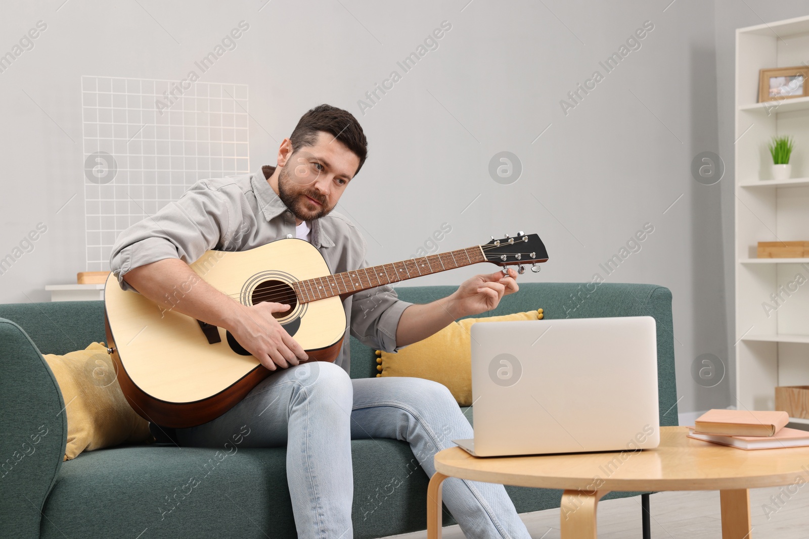 Photo of Relaxing hobby. Man playing guitar with online tutorial by laptop on sofa at home