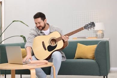 Photo of Relaxing hobby. Smiling man playing guitar with online tutorial by laptop on sofa at home