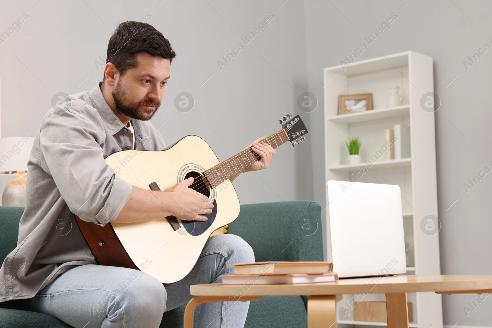 Photo of Relaxing hobby. Man playing guitar with online tutorial by laptop on sofa at home