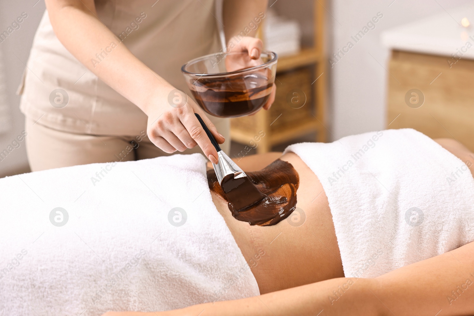 Photo of Chocolate body wrap. Spa worker applying mask onto woman's belly in salon, closeup