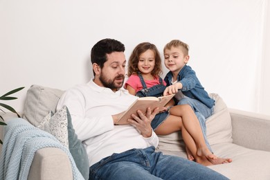 Photo of Overwhelmed father reading book to his children on sofa at home