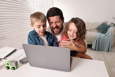 Photo of Naughty children disturbing their overwhelmed screaming father at table with laptop indoors