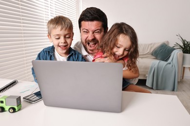 Photo of Naughty children disturbing their overwhelmed screaming father at table with laptop indoors