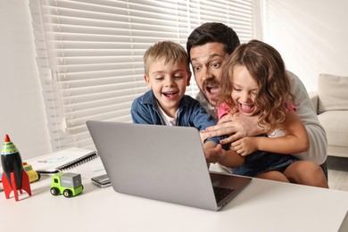 Photo of Naughty children disturbing their overwhelmed father at table with laptop indoors