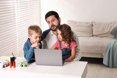 Photo of Naughty children disturbing their overwhelmed father at table with laptop indoors