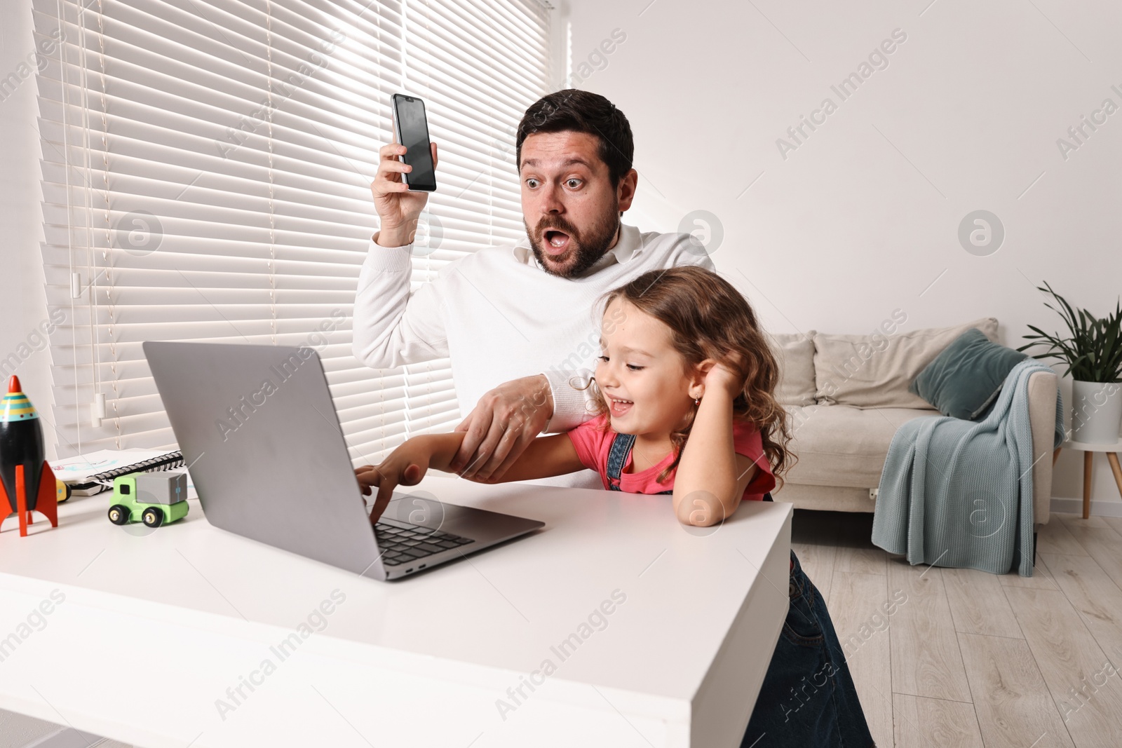 Photo of Naughty daughter disturbing her overwhelmed father at table with laptop indoors