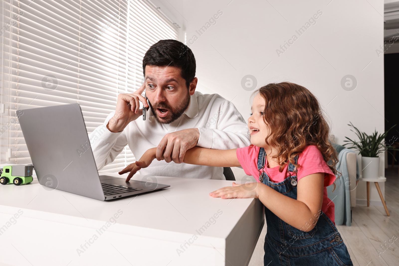 Photo of Naughty daughter disturbing her overwhelmed father while he talking on smartphone at table indoors