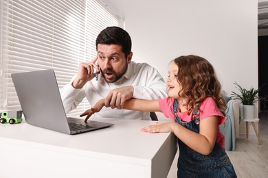 Photo of Naughty daughter disturbing her overwhelmed father while he talking on smartphone at table indoors