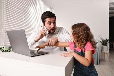 Photo of Naughty daughter disturbing her overwhelmed father while he talking on smartphone at table indoors