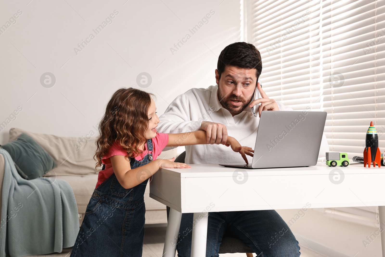 Photo of Naughty daughter disturbing her overwhelmed father while he talking on smartphone at table indoors