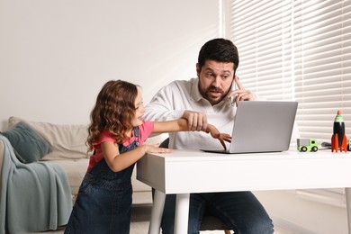 Photo of Naughty daughter disturbing her overwhelmed father while he talking on smartphone at table indoors