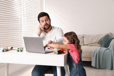 Photo of Naughty daughter disturbing her overwhelmed father while he talking on smartphone at table indoors