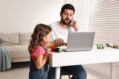 Photo of Naughty daughter disturbing her overwhelmed father while he talking on smartphone at table indoors