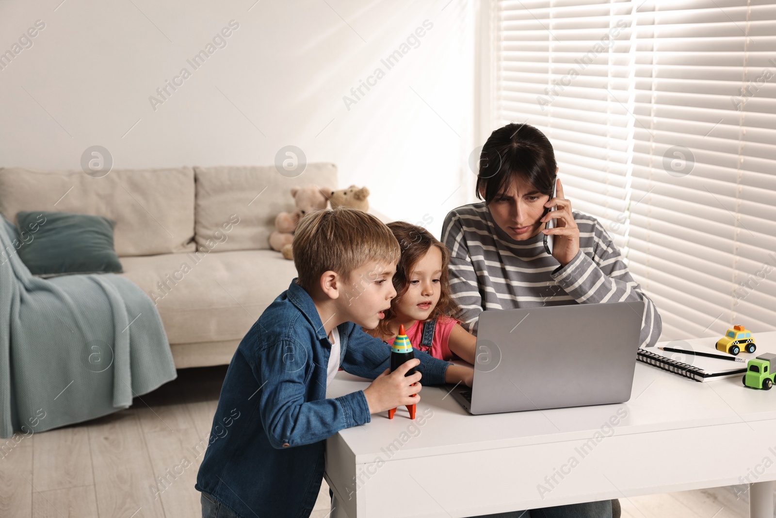 Photo of Naughty children disturbing overwhelmed mother while she talking on smartphone at table indoors