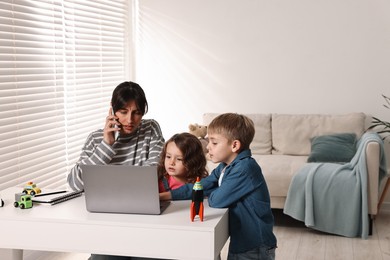 Photo of Naughty children disturbing overwhelmed mother while she talking on smartphone at table indoors