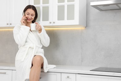 Photo of Beautiful woman with cup of drink talking on smartphone after spa procedure in kitchen