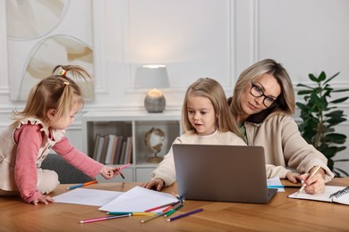 Photo of Work-family balance. Single mother talking by smartphone and her children drawing at wooden table indoors