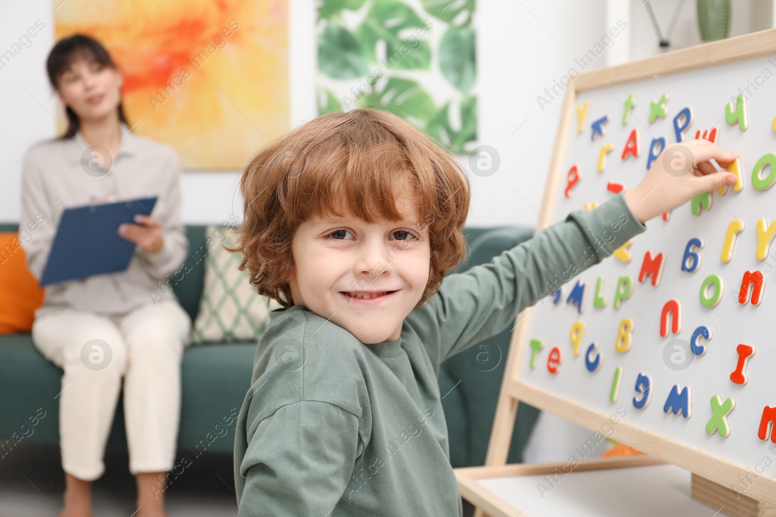 Photo of Boy assembling letters on magnetic board while psychologist taking notes indoors, selective focus