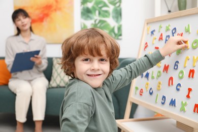 Photo of Boy assembling letters on magnetic board while psychologist taking notes indoors, selective focus