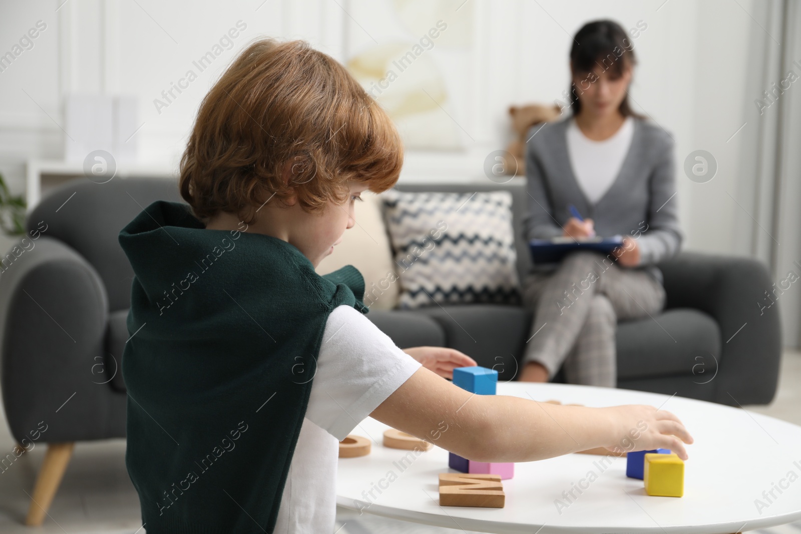 Photo of Boy playing with wooden cubes at table while psychologist taking notes, selective focus