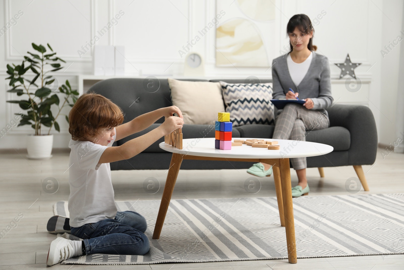 Photo of Boy playing with wooden letters at table while psychologist taking notes, selective focus