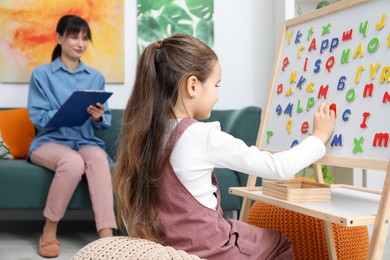 Photo of Girl assembling letters on magnetic board while psychologist taking notes indoors, selective focus