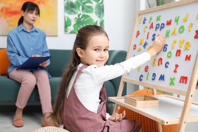 Photo of Girl assembling letters on magnetic board while psychologist taking notes indoors, selective focus