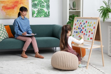 Photo of Girl assembling letters on magnetic board while psychologist taking notes indoors