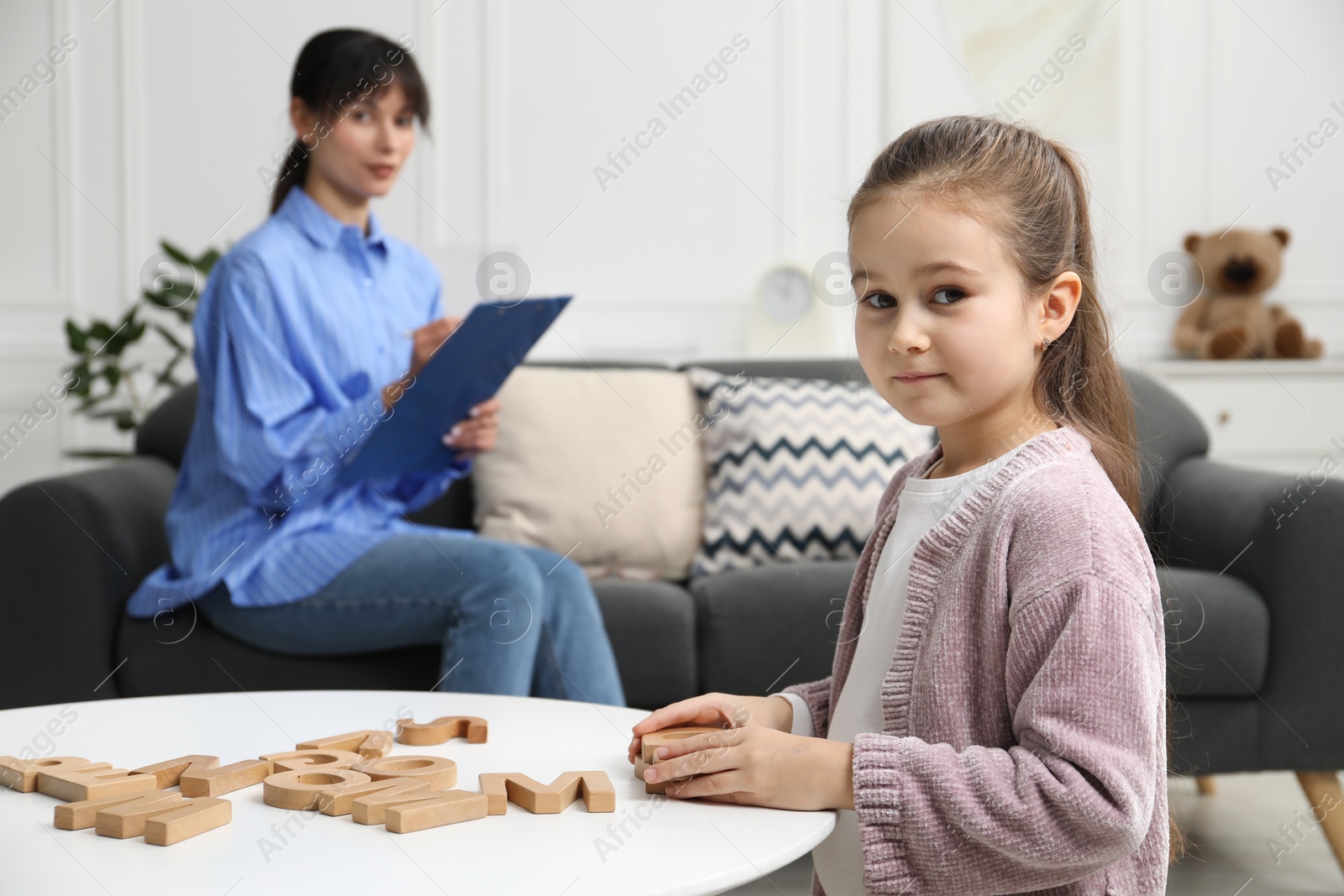 Photo of Girl playing with letters at table while psychologist taking notes, selective focus