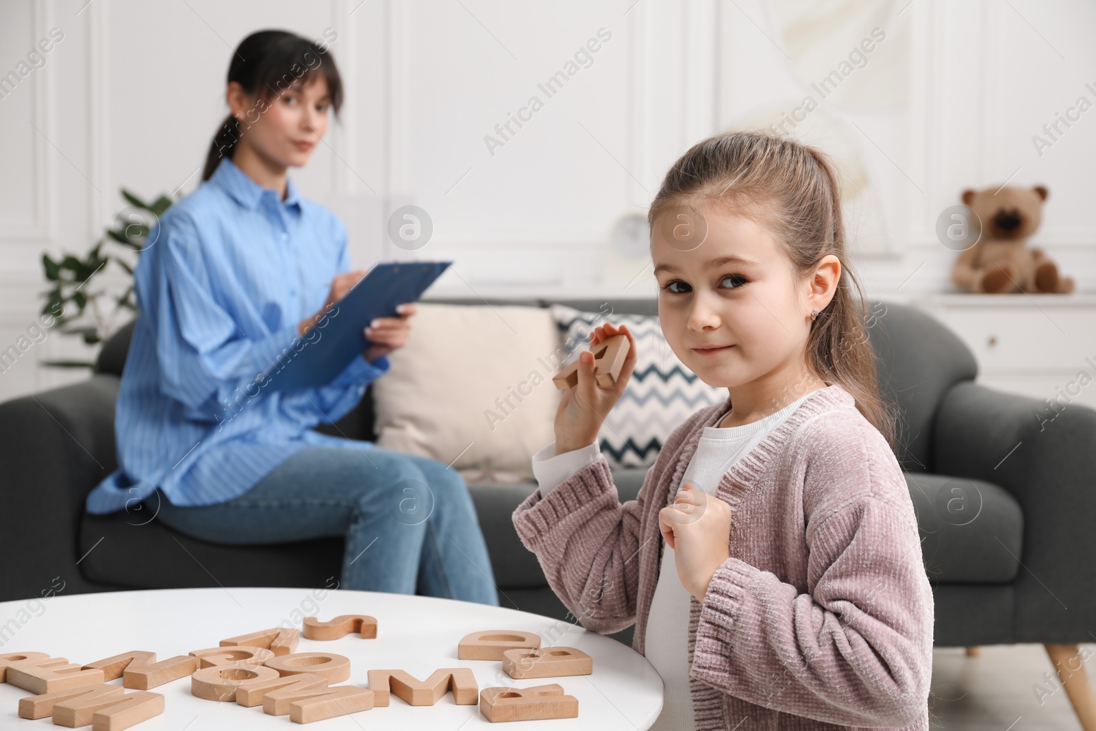 Photo of Girl playing with letters at table while psychologist taking notes, selective focus