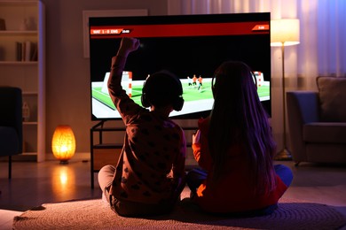 Photo of Little kids in headphones playing video game indoors at night, back view