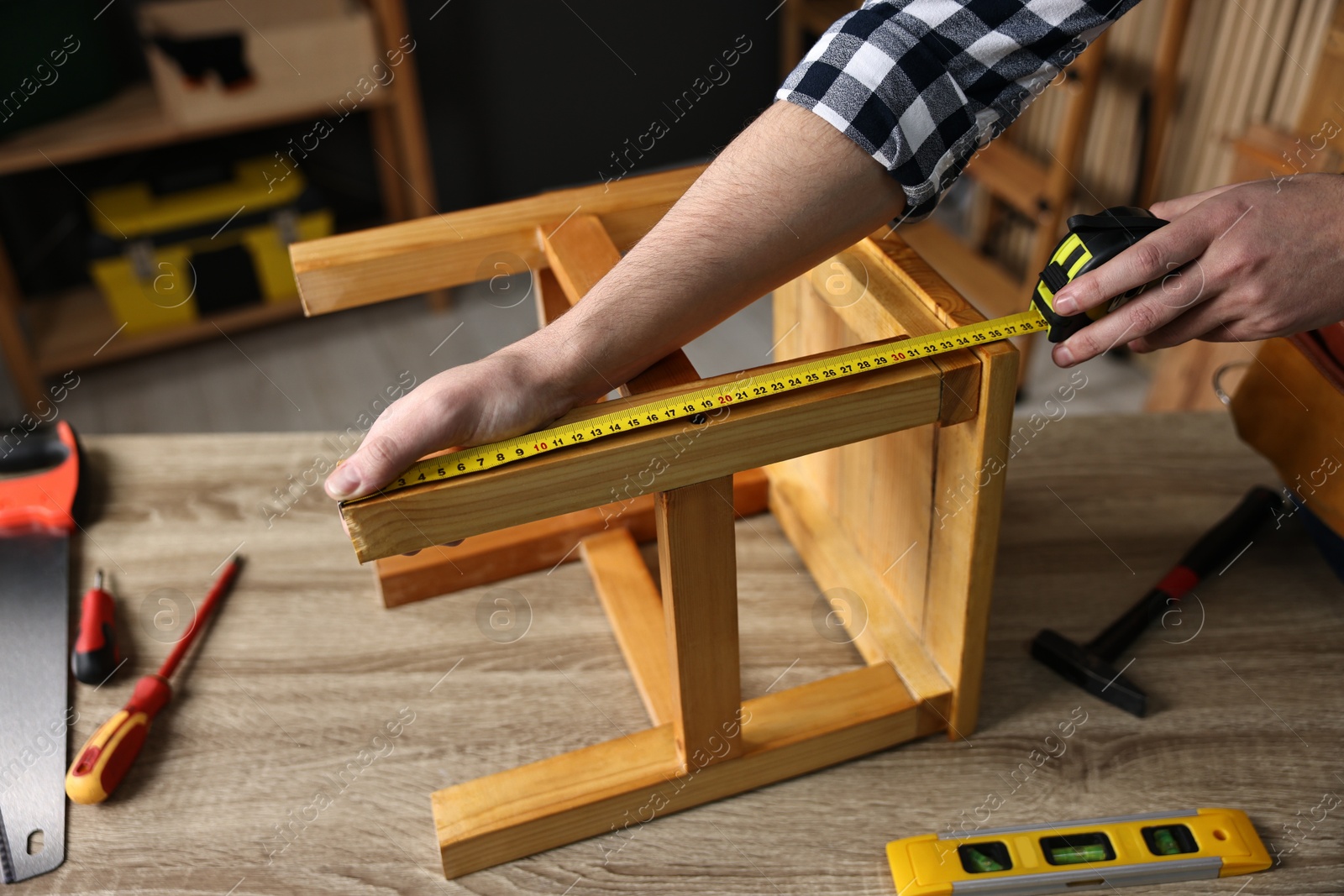 Photo of Repairman measuring wooden stool at table in workshop, closeup