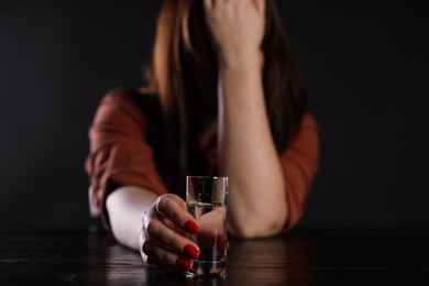 Photo of Alcohol addiction. Miserable woman with vodka at table in dark, selective focus