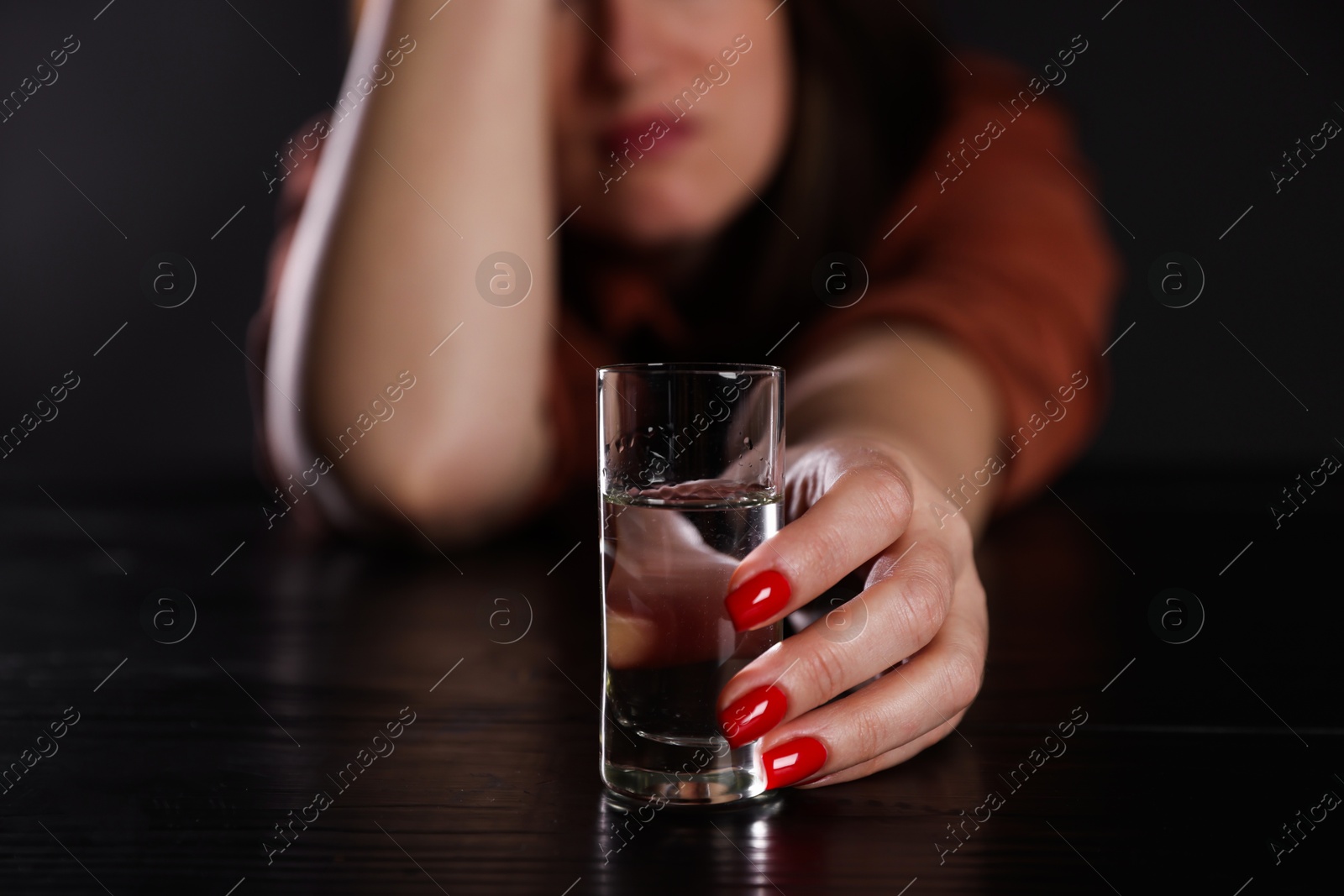 Photo of Alcohol addiction. Miserable woman with vodka at table in dark, closeup